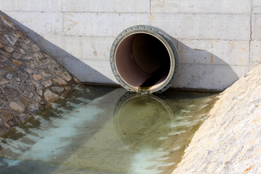 Large diameter plastic pipe storm drain drainage outlet covered with concrete leading to drain canal surrounded with stone tiles and water on cold sunny winter day